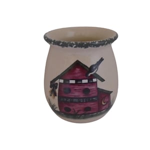 May include: A ceramic mug with a cream-colored background and a painted design of a red birdhouse with a black bird perched on top. The birdhouse has a green roof and white trim. The mug has a green rim.