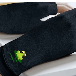K&ouml;nnte beinhalten: Paar schwarze fingerlose Handschuhe mit einem gr&uuml;nen Froschdesign auf jedem Handschuh. Die Handschuhe sind aus einem weichen, atmungsaktiven Stoff gefertigt und sollen helfen, Handm&uuml;digkeit zu reduzieren.
