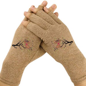 May include: Beige fingerless gloves with a cartoon cat design. The cat wears glasses and a pink bow tie, perched on a black branch with green leaves. The gloves are made of a soft, textured material.