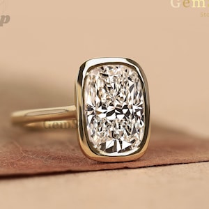 May include: A gold ring with a large, rectangular, clear gemstone. The gemstone is set in a gold bezel and has a brilliant cut. The ring is on a brown surface. The GemShopStore logo is in the background.