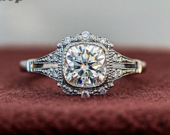 May include: A silver engagement ring featuring a large, square-cut diamond. The diamond is encircled by smaller accent stones, and the ring rests on a dark red velvet surface. The ring showcases detailed craftsmanship.