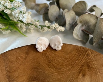 Puede incluir: Un par de pendientes de botón de calavera blanca. Los pendientes están hechos de un material blanco liso y tienen un diseño de calavera ligeramente elevado.