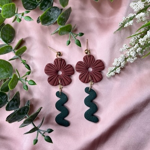 May include: Handmade earrings with maroon flower tops and gold hooks. Dark green, wavy shapes hang below the flowers. The earrings are set against a pink backdrop, with green and white floral decorations. The earrings are a unique accessory.