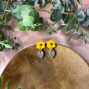 May include: A pair of yellow flower earrings with grey leaf pendants. The earrings are set on a wooden plate, with green leafy branches in the background. A bright and cheerful accessory.