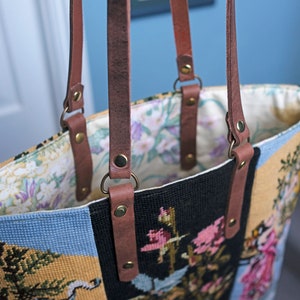 May include: A brown leather tote bag with a floral tapestry design. The bag has two brown leather straps with brass buckles and rivets.