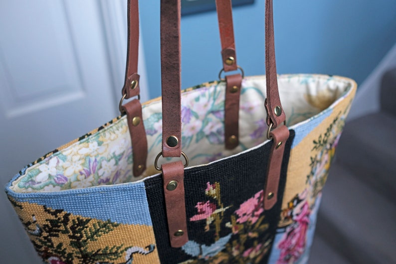 May include: A brown leather tote bag with a floral tapestry design. The bag has two brown leather straps with brass buckles and rivets.