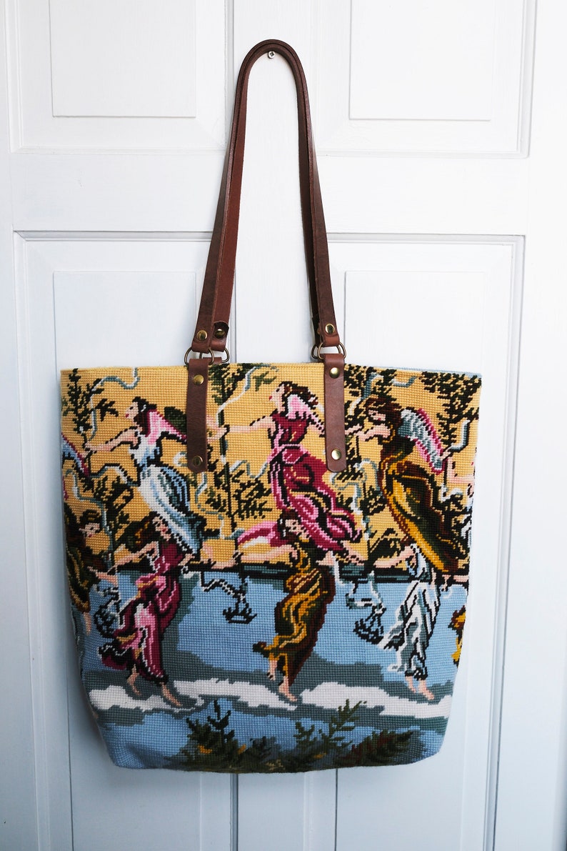 May include: A yellow and blue tapestry tote bag with brown leather straps. The tapestry features a colorful scene of people dancing in a garden.
