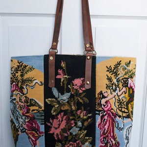 May include: A black, yellow, and blue tapestry tote bag with brown leather straps and gold hardware. The tapestry features a floral design with a scene of people in a garden.