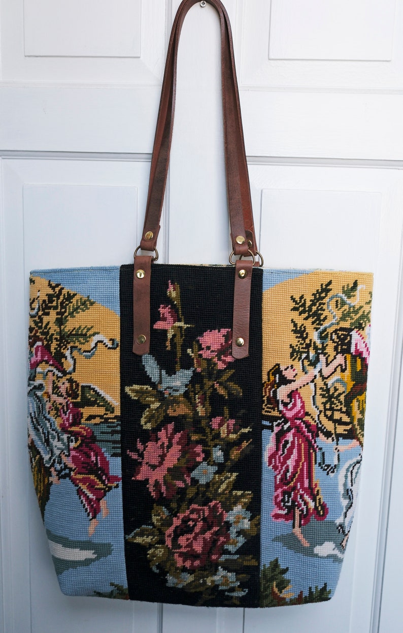 May include: A black, yellow, and blue tapestry tote bag with brown leather straps and gold hardware. The tapestry features a floral design with a scene of people in a garden.