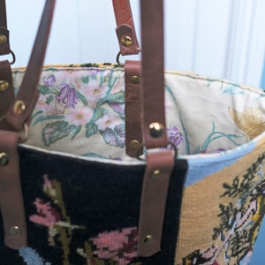 May include: A brown leather tote bag with a floral patterned lining. The bag is made of a tapestry fabric with a floral design in shades of blue, yellow, and black.