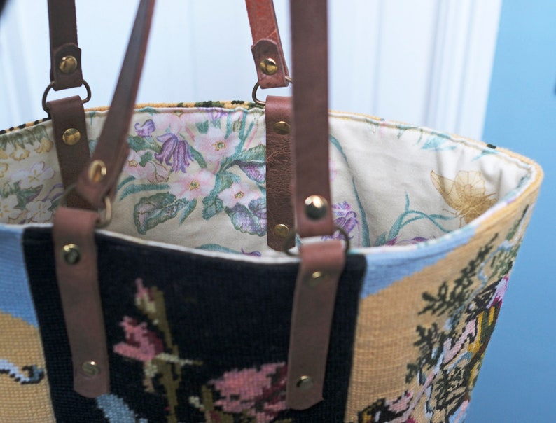May include: A brown leather tote bag with a floral patterned lining. The bag is made of a tapestry fabric with a floral design in shades of blue, yellow, and black.