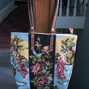 May include: A tapestry tote bag with a floral and figural design. The bag has brown leather straps and gold hardware.