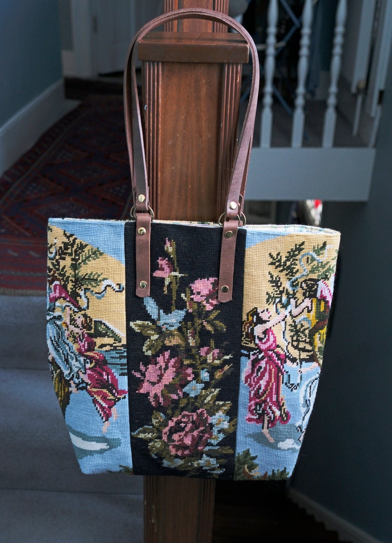 May include: A tapestry tote bag with a floral and figural design. The bag has brown leather straps and gold hardware.