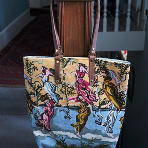 May include: A brown leather tote bag with a tapestry design featuring a colorful scene of people dancing in a garden. The bag has a brown leather strap and gold metal accents.