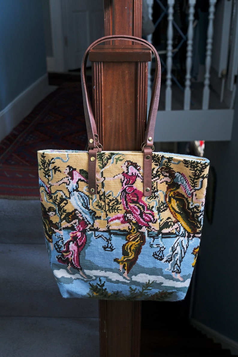 May include: A brown leather tote bag with a tapestry design featuring a colorful scene of people dancing in a garden. The bag has a brown leather strap and gold metal accents.