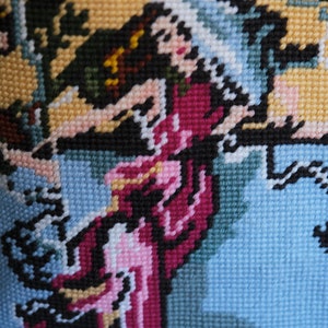 May include: A close-up of a colorful needlepoint embroidery depicting a woman in a pink and purple dress with a blue background.