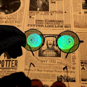 Peut inclure: Lunettes rondes avec des montures fines en métal noir et des verres vert vif. Chaque verre présente un graphique détaillé et lumineux d'un globe oculaire humain avec des veines visibles et un iris central, créant un effet visuel saisissant.
