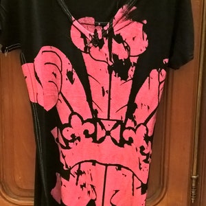 Può includere: Una t-shirt nera a maniche corte con una grande stampa grafica rosa acceso in stile consumato che raffigura l'emblema delle piume del Principe di Galles sul davanti. La maglia presenta uno scollo a V con cuciture bianche visibili sui bordi.