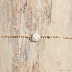 May include: A delicate gold chain bracelet featuring a white and tan shell bead. The bead is the focal point, with a spiral design. The bracelet is displayed against a light brown wooden background.