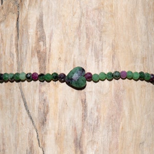 Può includere: Un braccialetto di perline con un ciondolo a forma di cuore in pietra preziosa verde e viola, composto da perline verdi, viola e nere.