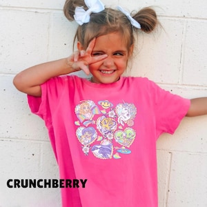 May include: A bright pink t-shirt with a colorful graphic design featuring various cartoon characters, including a unicorn, alien, and tiger. The word "CRUNCHBERRY" is printed at the bottom of the shirt.