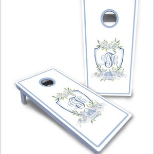 May include: Two white cornhole boards with light blue borders and circular holes. Each board features a central crest design with blue floral watercolor illustrations, a decorative monogram, and the printed text June 22, 2024.