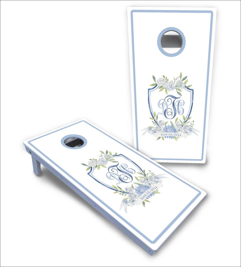 May include: Two white cornhole boards with light blue borders and circular holes. Each board features a central crest design with blue floral watercolor illustrations, a decorative monogram, and the printed text June 22, 2024.