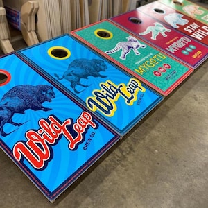 May include: A series of rectangular cornhole boards featuring vibrant graphic designs. The boards display illustrations of bison, leopards, and bears, alongside the text Wild Leap Brew Co., Mygotu, and Stay Wild, with circular holes at the top.
