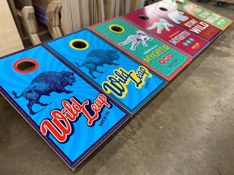 May include: A series of rectangular cornhole boards featuring vibrant graphic designs. The boards display illustrations of bison, leopards, and bears, alongside the text Wild Leap Brew Co., Mygotu, and Stay Wild, with circular holes at the top.
