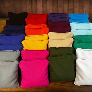 May include: A collection of square fabric bean bags arranged in a grid, featuring a variety of solid colors including black, red, purple, blue, orange, yellow, beige, green, grey, pink, olive, and white, each with a visible stitched seam.