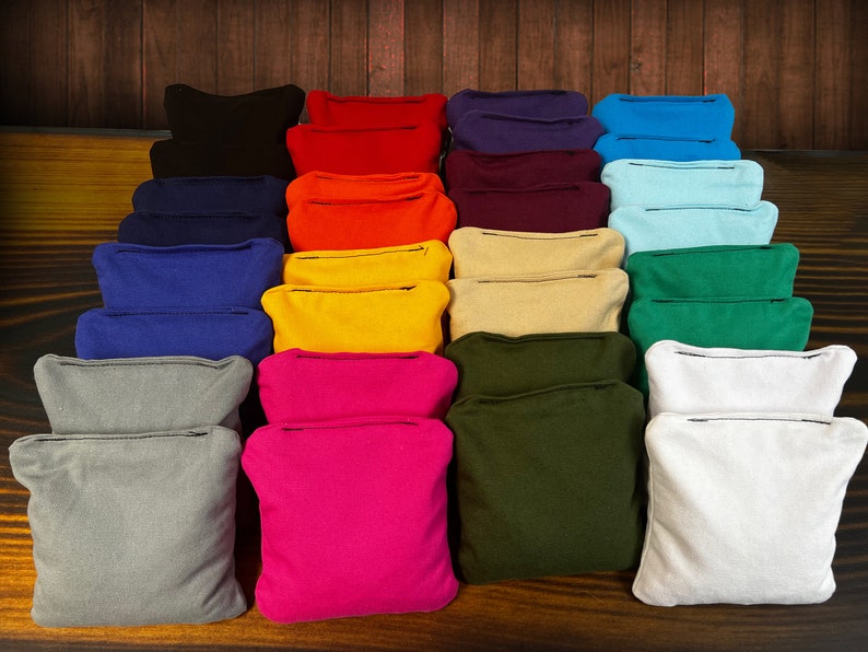 May include: A collection of square fabric bean bags arranged in a grid, featuring a variety of solid colors including black, red, purple, blue, orange, yellow, beige, green, grey, pink, olive, and white, each with a visible stitched seam.