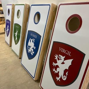 May include: Four wooden cornhole boards with white surfaces and natural wood sides. Each board features a colored shield graphic with a mythical creature and a name: Virgil in red, Homer in blue, Dante in green, and Milton in purple. Each board has a circular hole.
