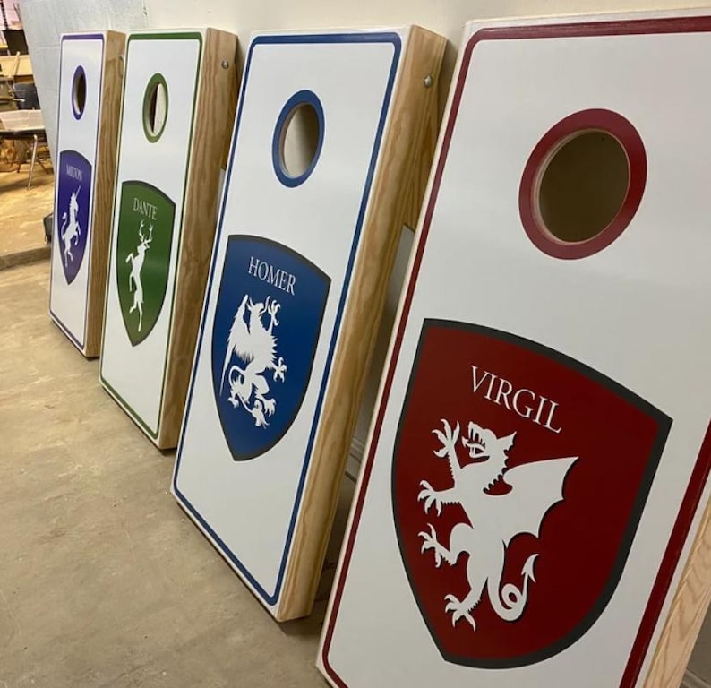 May include: Four wooden cornhole boards with white surfaces and natural wood sides. Each board features a colored shield graphic with a mythical creature and a name: Virgil in red, Homer in blue, Dante in green, and Milton in purple. Each board has a circular hole.