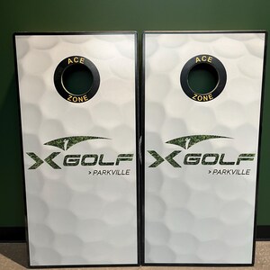 May include: Two rectangular cornhole boards featuring a white golf ball dimple pattern. Each board has a circular hole at the top with black trim labeled ACE ZONE. The center displays the X-GOLF logo with a golfer graphic and the text > PARKVILLE.