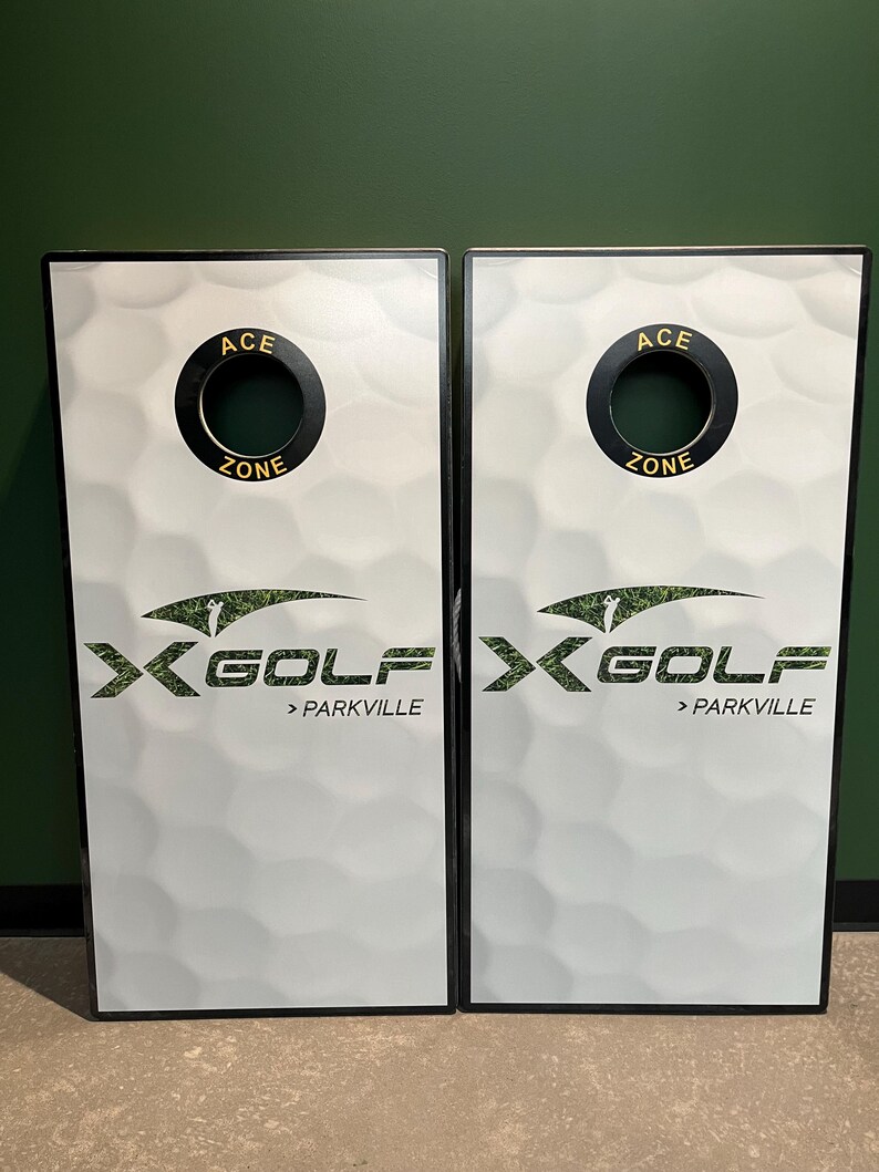 May include: Two rectangular cornhole boards featuring a white golf ball dimple pattern. Each board has a circular hole at the top with black trim labeled ACE ZONE. The center displays the X-GOLF logo with a golfer graphic and the text > PARKVILLE.