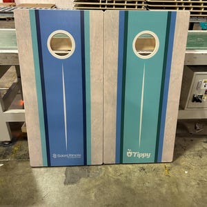 May include: Two rectangular wooden cornhole boards featuring a light wood grain finish and vertical stripes in shades of blue and teal. Each board has a circular hole at the top. The left board displays the text SalonUltimate and the right board says Tippy.