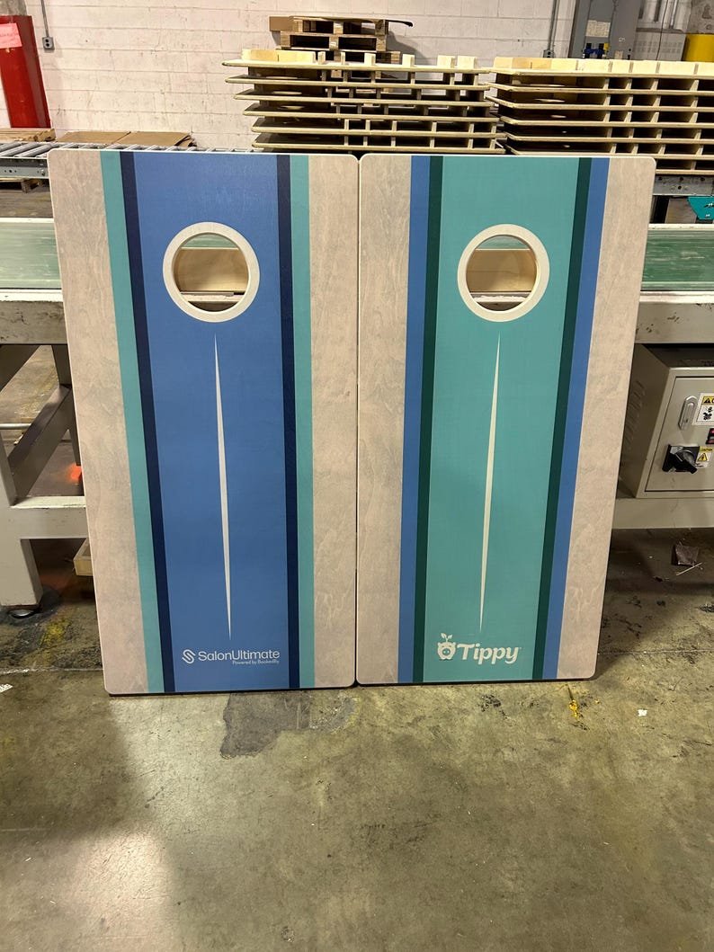 May include: Two rectangular wooden cornhole boards featuring a light wood grain finish and vertical stripes in shades of blue and teal. Each board has a circular hole at the top. The left board displays the text SalonUltimate and the right board says Tippy.