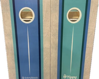 May include: Two cornhole boards with blue and teal stripes and a central hole. One board has the text "SalonUltimate" and the other has "Tippy". The boards are made of wood and have a light-colored wood grain.