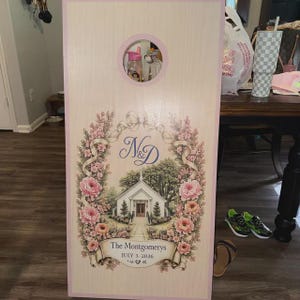May include: A rectangular wooden cornhole board with a light wood grain finish and a pink border. The board features a circular hole at the top and a central illustration of a white church surrounded by pink floral garlands, the letters N&D, and the text The Montgomerys JULY 3, 2026.