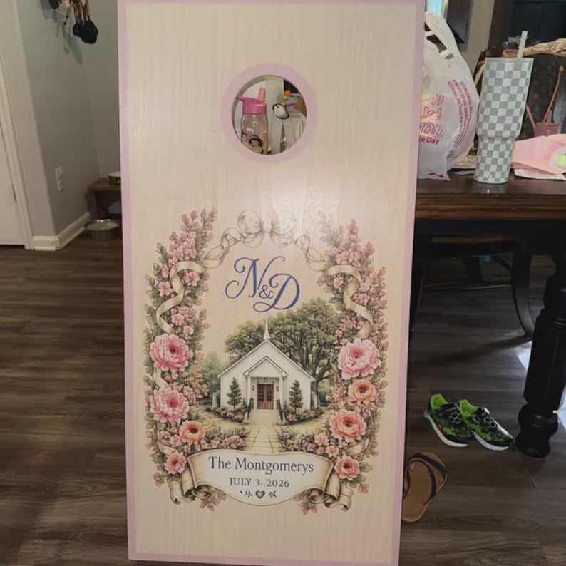May include: A rectangular wooden cornhole board with a light wood grain finish and a pink border. The board features a circular hole at the top and a central illustration of a white church surrounded by pink floral garlands, the letters N&D, and the text The Montgomerys JULY 3, 2026.