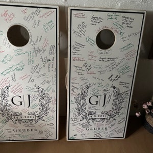 Custom cornhole, cornhole boards, customboards, cornhole score board, backyard games, custom logo boards, monogram corn hole, cornhole board, corn hole boards, custom game board, groomsman gift, monogram cornhole, wedding decal. Two white wooden boards with black trim feature a wreath design with the initials "GJ" and the date "9.9.2023." Covered with handwritten messages and signatures, they create a unique wedding keepsake and personalized cornhole set.
