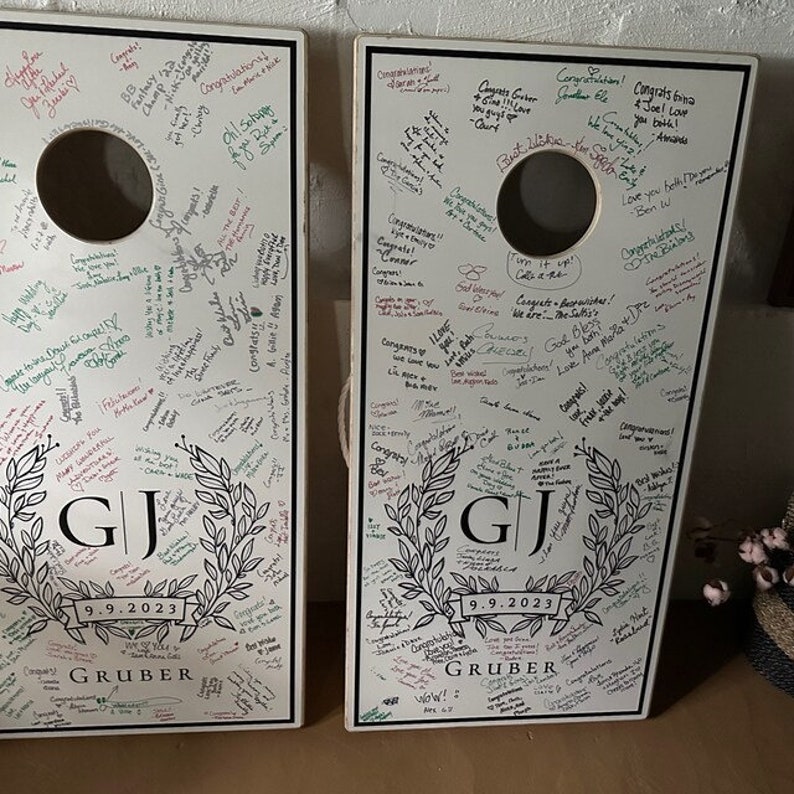Custom cornhole, cornhole boards, customboards, cornhole score board, backyard games, custom logo boards, monogram corn hole, cornhole board, corn hole boards, custom game board, groomsman gift, monogram cornhole, wedding decal. Two white wooden boards with black trim feature a wreath design with the initials "GJ" and the date "9.9.2023." Covered with handwritten messages and signatures, they create a unique wedding keepsake and personalized cornhole set.