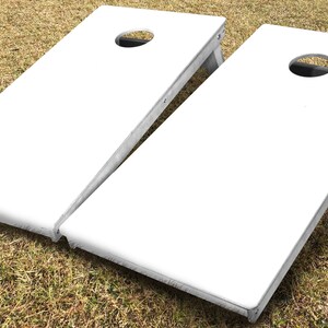 May include: Two white wooden cornhole boards with a circular hole in the centre of each board. The boards are on a grassy surface.