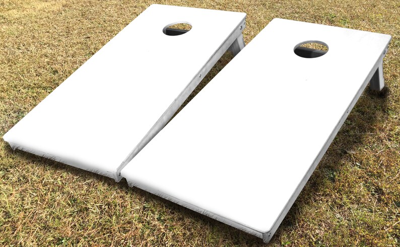 May include: Two white wooden cornhole boards with a circular hole in the centre of each board. The boards are on a grassy surface.