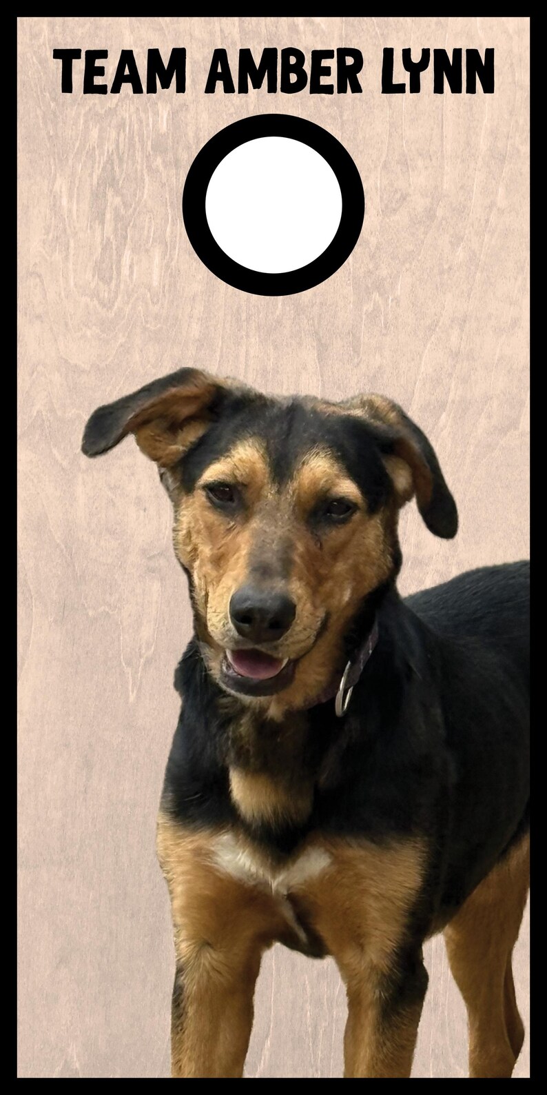 May include: A cornhole board with a light wood grain finish featuring the text TEAM AMBER LYNN in black capital letters above a circular hole with a black border and a photograph of a black and tan dog with floppy ears and a collar.