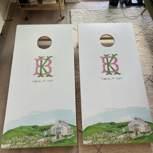 May include: Two white cornhole boards featuring a circular hole at the top. Each board displays a pink and green monogram of the letters B and K, followed by the text September 22, 2024. The bottom shows a painted landscape of a barn and flowers.