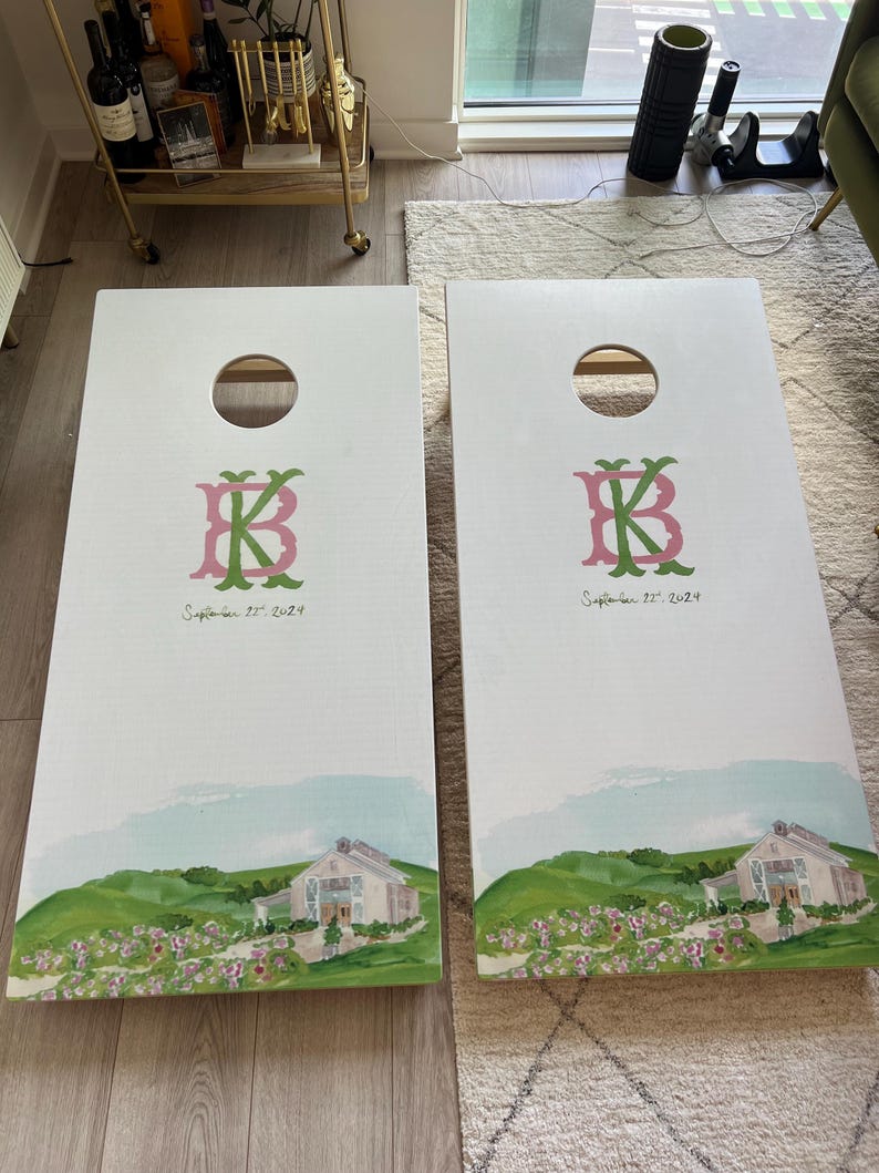 May include: Two white cornhole boards featuring a circular hole at the top. Each board displays a pink and green monogram of the letters B and K, followed by the text September 22, 2024. The bottom shows a painted landscape of a barn and flowers.