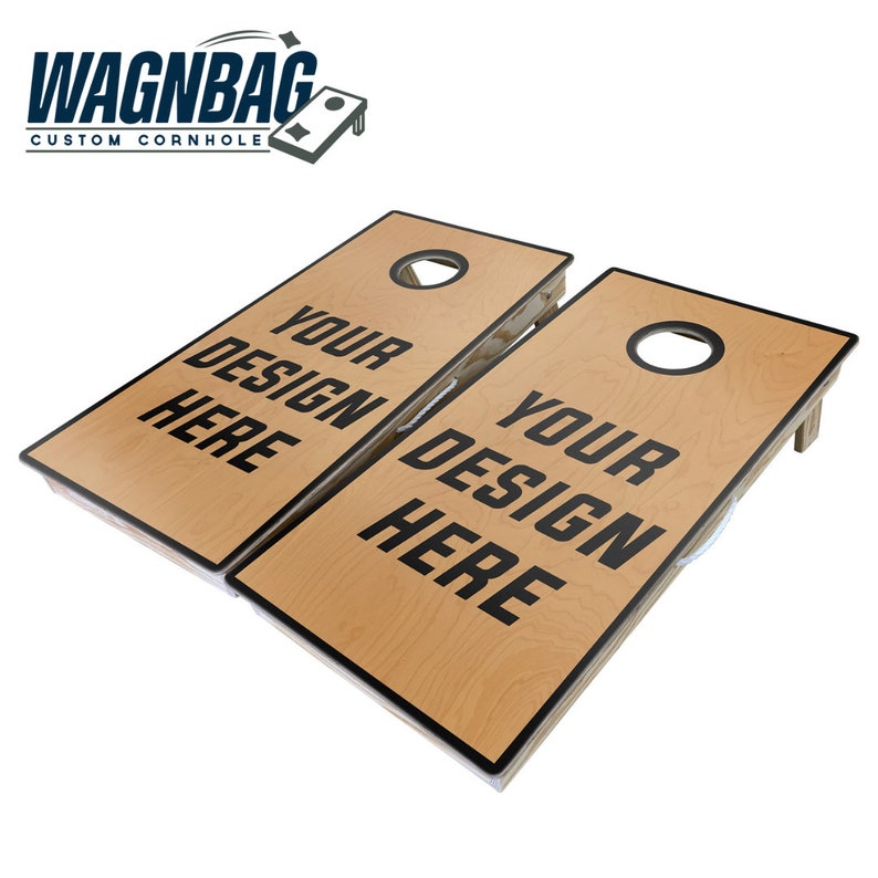 May include: Two light-colored wooden cornhole boards with black trim and circular holes at the top. Each board features the black text "YOUR DESIGN HERE" printed in the center. The boards are angled and include folding legs for support.
