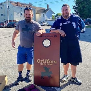 May include: A rectangular cornhole board with a dark red surface, a circular hole at the top, and black graphics featuring a winged pig. The text reads "FOLLOW US ON FACEBOOK" and "Griffins FOOD FOR THE SOUL" in a light-colored, sans-serif font.
