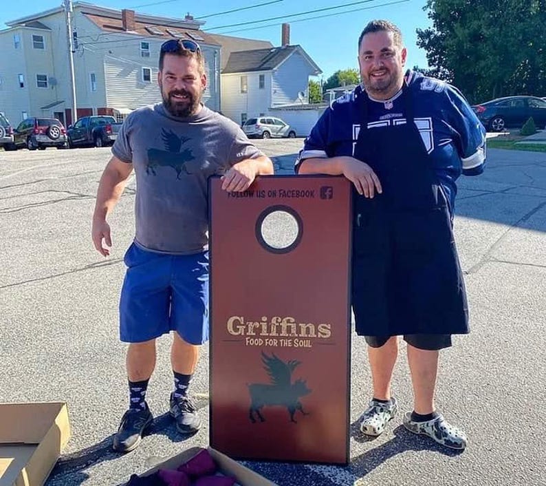 May include: A rectangular cornhole board with a dark red surface, a circular hole at the top, and black graphics featuring a winged pig. The text reads "FOLLOW US ON FACEBOOK" and "Griffins FOOD FOR THE SOUL" in a light-colored, sans-serif font.